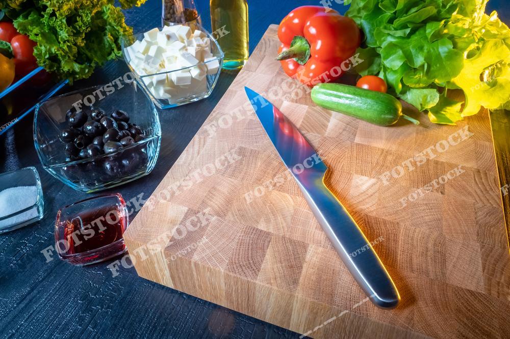 На фотографии изображена деревянная кухонная доска с ножом, рядом расположены свежие овощи: болгарский перец, огурцы, зелень, а также ингредиенты в стеклянных мисках, такие как маслины, сыр фета и специи. Композиция подчёркивает процесс подготовки натуральных продуктов для приготовления блюда.